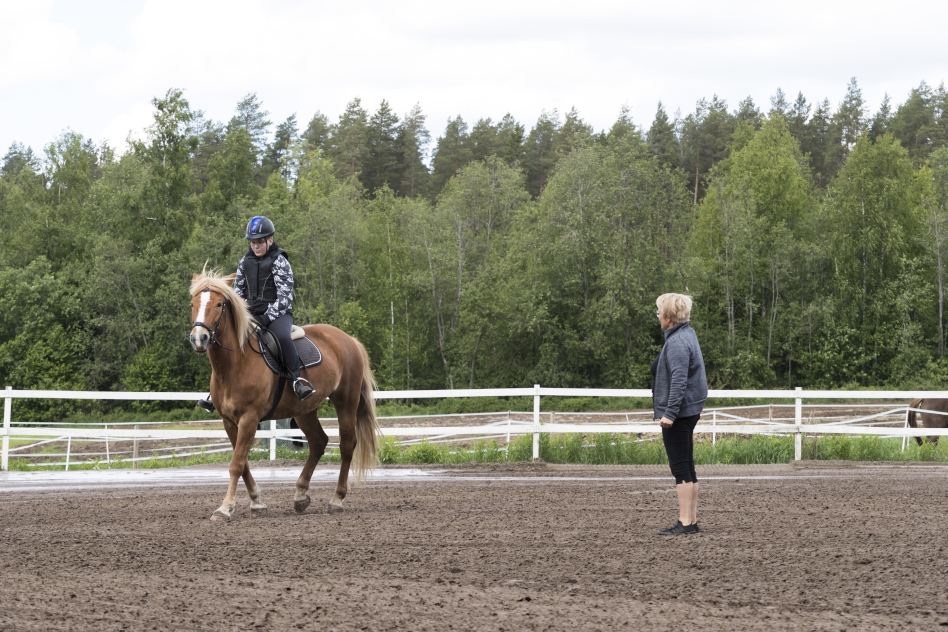 Nainen ratsastaa vaaleanruskealla hevosella, ohjaaja seisoo lähellä